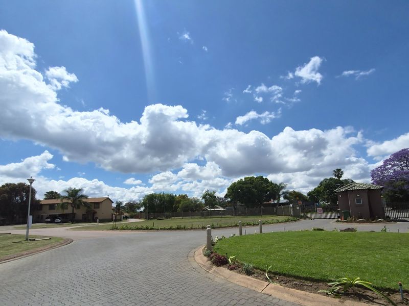 A view of the residential estate entrance featuring a paved driveway, landscaped green space, and a security gatehouse under a clear blue sky.