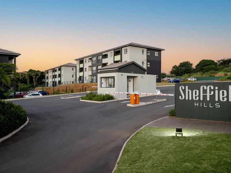 The entrance to Sheffield Hills apartment complex featuring a secure boom gate, guardhouse, and well-maintained paved driveway and landscaped grounds.