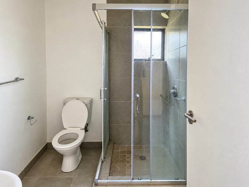A functional bathroom featuring a glass-enclosed shower with tiled walls and a standard toilet, offering a clean and simple layout.