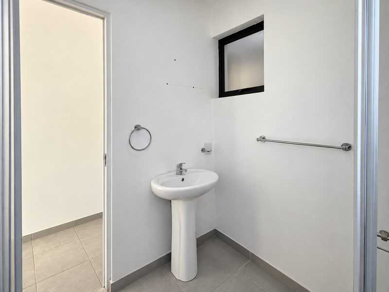 A clean and minimalist bathroom featuring a pedestal basin, chrome fixtures, and neutral tiled flooring with a frosted window for natural light.