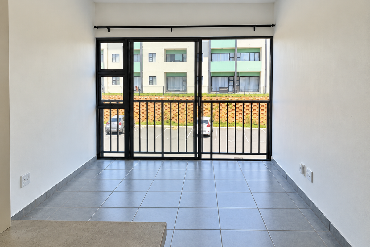 A bright, empty living area featuring neutral tiled flooring and a large sliding glass door with a security gate, offering views of the parking area.