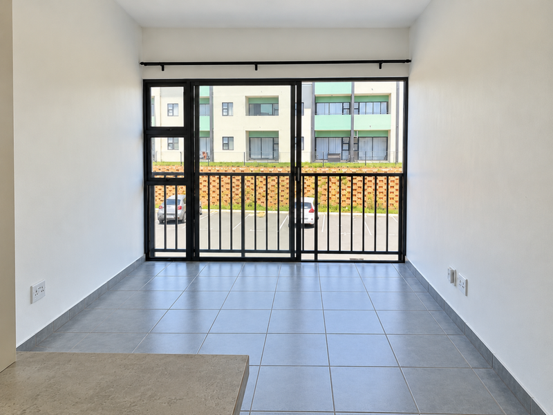 A bright, empty living area featuring neutral tiled flooring and a large sliding glass door with a security gate, offering views of the parking area.