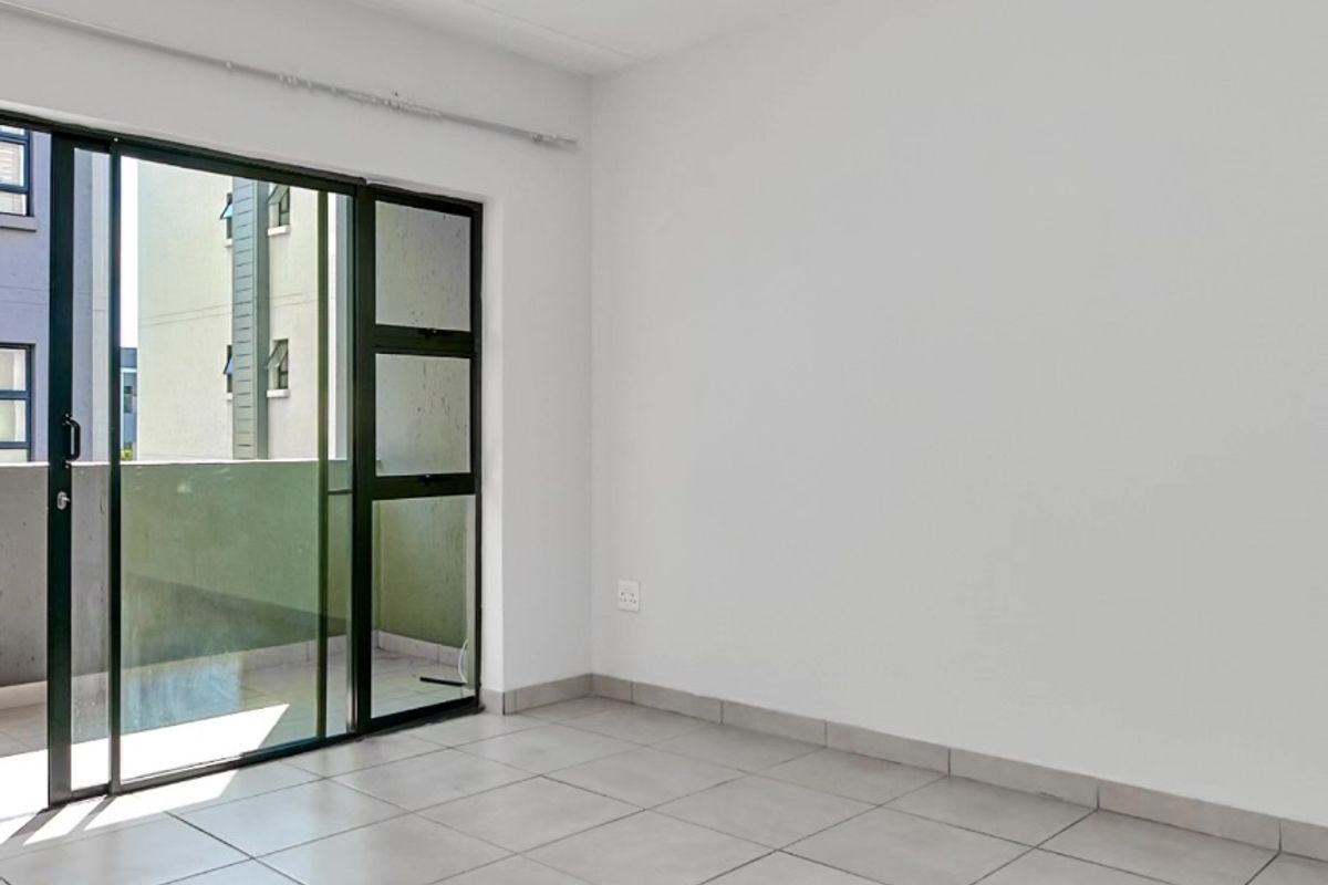 A bright, empty living area featuring neutral tiled flooring and a large sliding glass door providing access to an outdoor balcony.