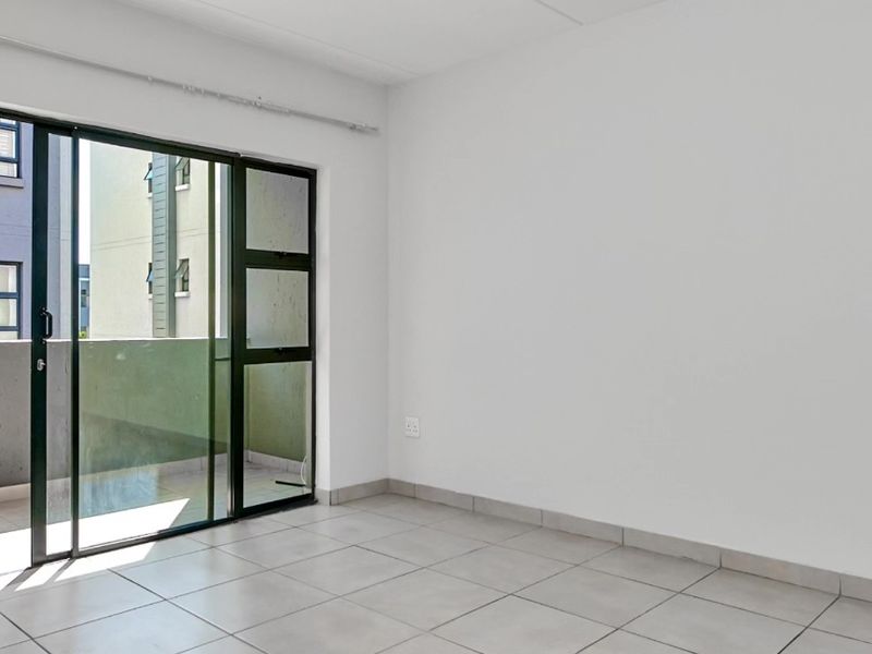 A bright, empty living area featuring neutral tiled flooring and a large sliding glass door providing access to an outdoor balcony.