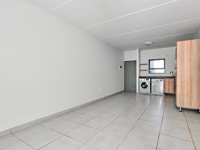 An open-plan living and kitchen area featuring neutral tiled flooring, clean white walls, and a functional kitchenette with built-in laundry appliance connections.