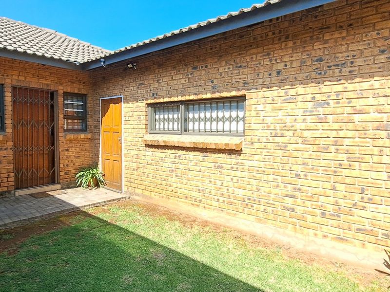 Rear exterior view of a face brick townhouse featuring a tiled roof, security gates, paved walkway, and a small private lawn area.