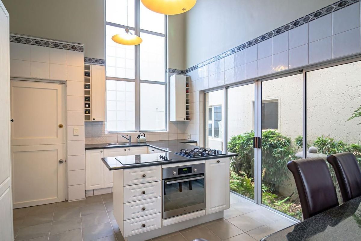 A bright kitchen featuring double-height ceilings, white cabinetry, granite countertops, a central island with a gas hob and oven, and sliding doors to a garden.
