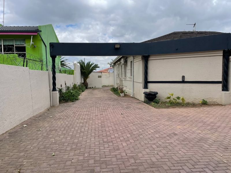 A paved driveway leading to a commercial property entrance, featuring a covered structure, perimeter walling with security spikes, and established garden beds.