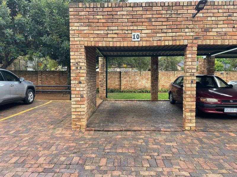 A covered brick carport structure with paved flooring, providing dedicated parking space within a secure complex setting.