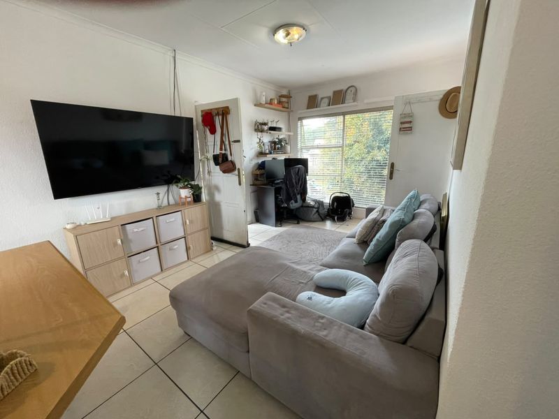A compact living room featuring tiled flooring, a ceiling light fixture, and a window with blinds, offering a functional space for relaxation.