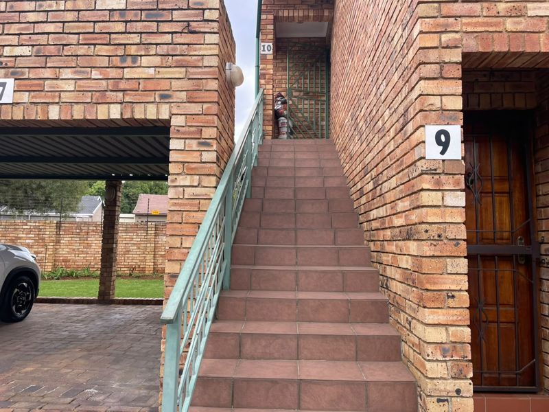 Exterior view of a townhouse entrance featuring a face brick facade, tiled staircase with metal balustrade, and an adjacent covered carport.