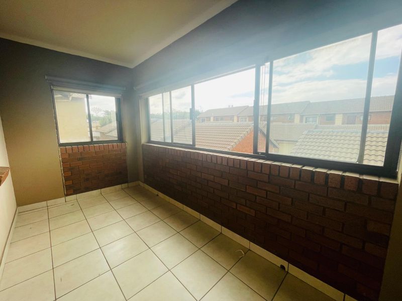 An enclosed balcony or sunroom featuring tiled flooring, brick wall accents, and large sliding windows offering views of the surrounding townhouse complex.