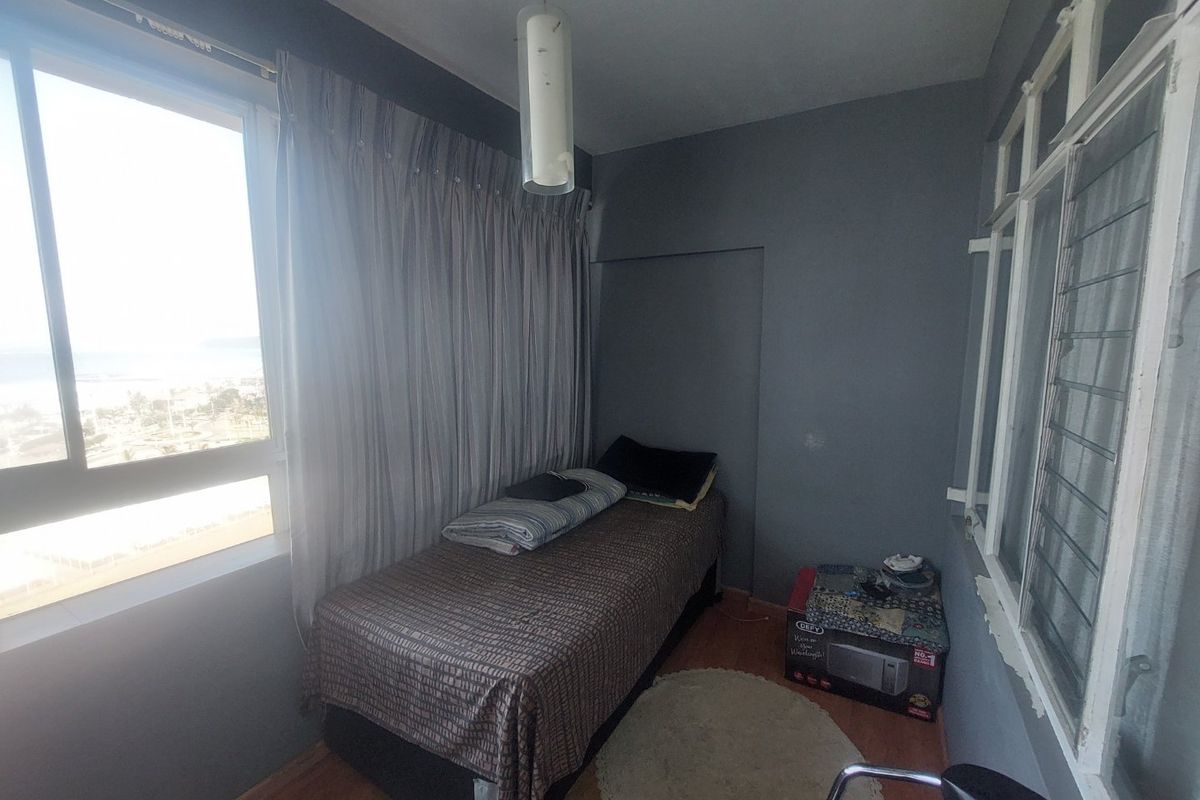 A compact bedroom featuring laminate flooring, a large sliding window with natural light, and additional louvered windows for ventilation.