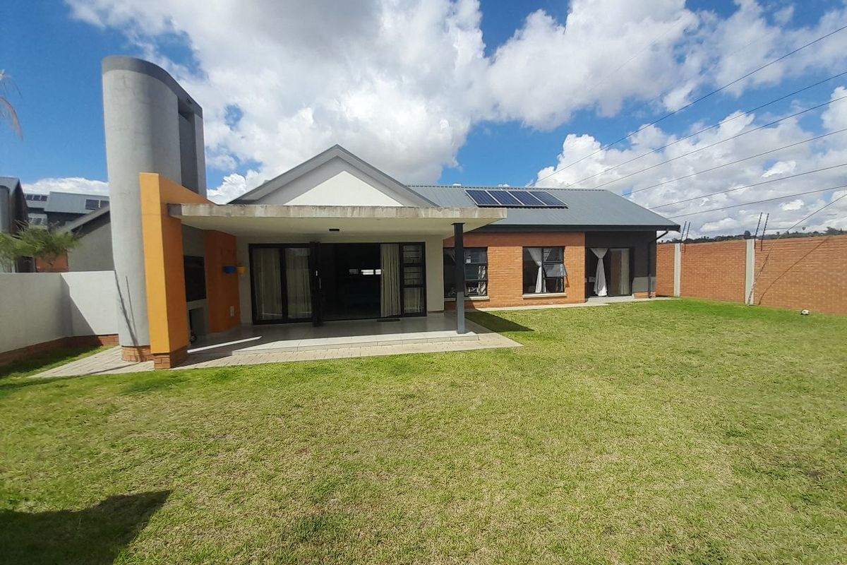 Rear view of the house featuring a covered patio with a built-in braai, solar panels, and a spacious garden enclosed by a brick boundary wall.