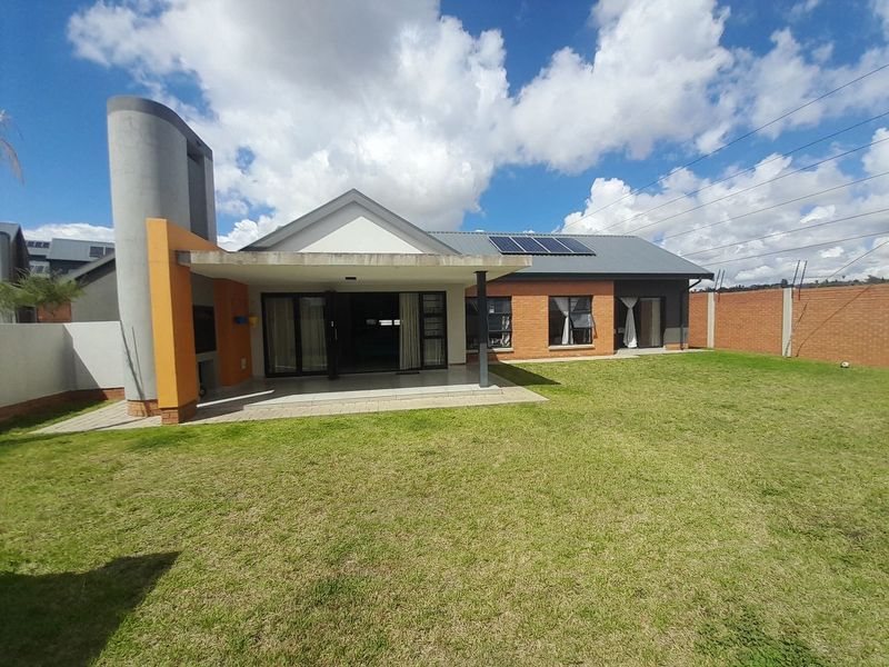 Rear view of the house featuring a covered patio with a built-in braai, solar panels, and a spacious garden enclosed by a brick boundary wall.