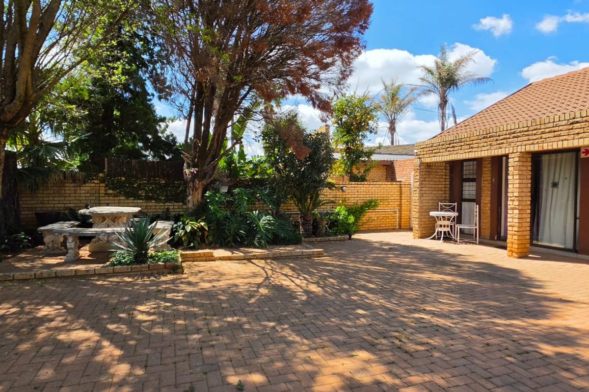 A spacious rear courtyard featuring brick paving, a covered patio area, and established garden landscaping with a facebrick building exterior.