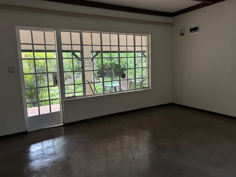 An empty living room featuring polished concrete floors, exposed ceiling beams, and large grid-pattern windows providing natural light and access to the outdoor patio area.
