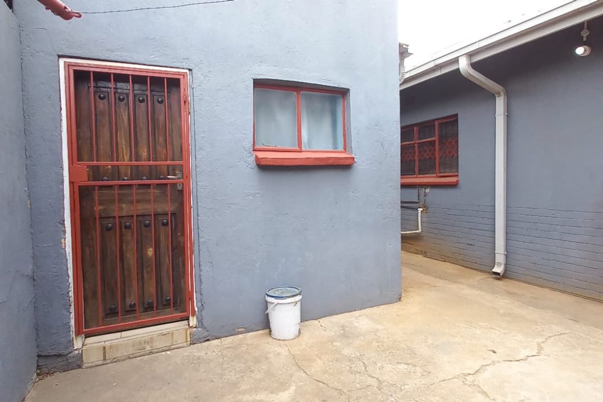 Rear exterior area featuring concrete paving, security gate, and painted brick walls with visible wear on window frames and paving.