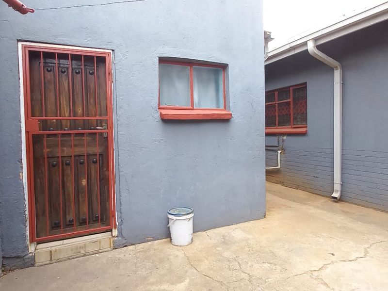 Rear exterior area featuring concrete paving, security gate, and painted brick walls with visible wear on window frames and paving.