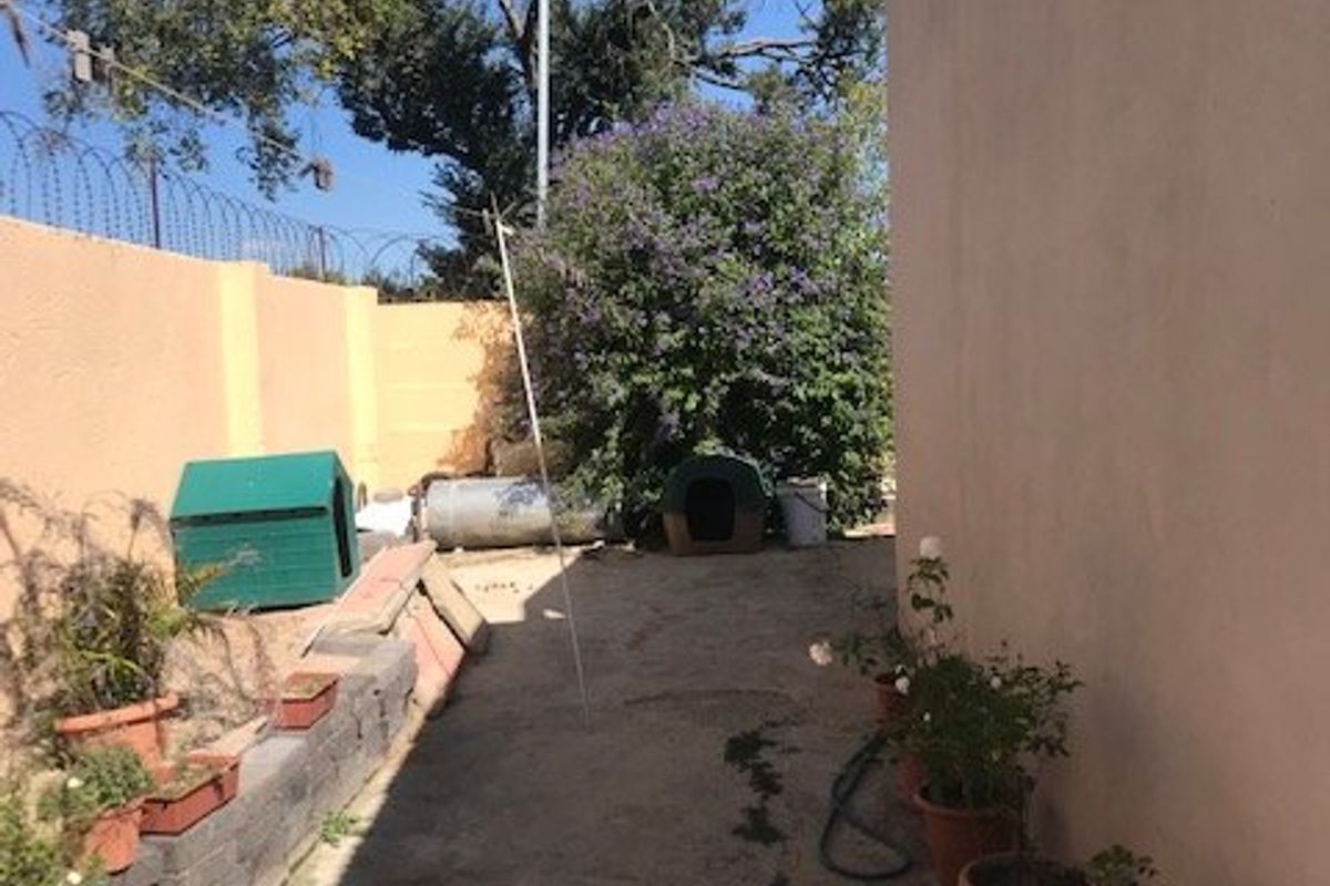A narrow rear exterior area featuring concrete paving, a perimeter wall with security fencing, and a small retaining wall.