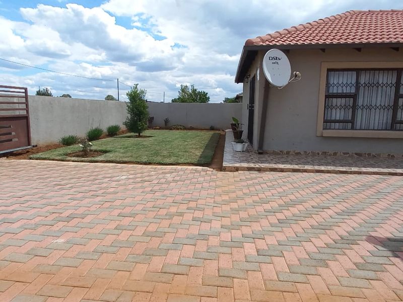 A well-maintained rear exterior featuring a paved driveway, a neat lawn, boundary walls, and a secure home facade with tiled roofing.
