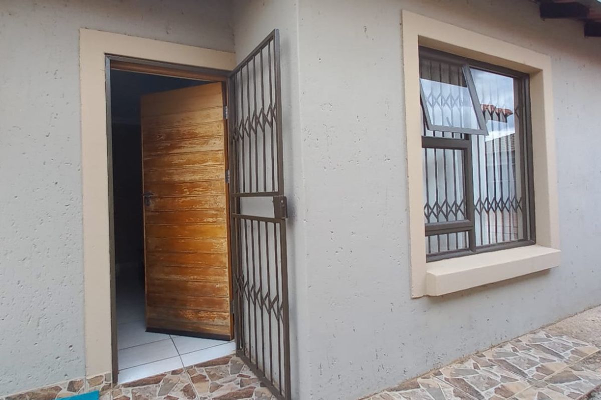 Rear exterior view of the house featuring a paved patio, security gate, wooden entrance door, and aluminium window frames with security bars.