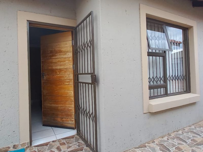 Rear exterior view of the house featuring a paved patio, security gate, wooden entrance door, and aluminium window frames with security bars.