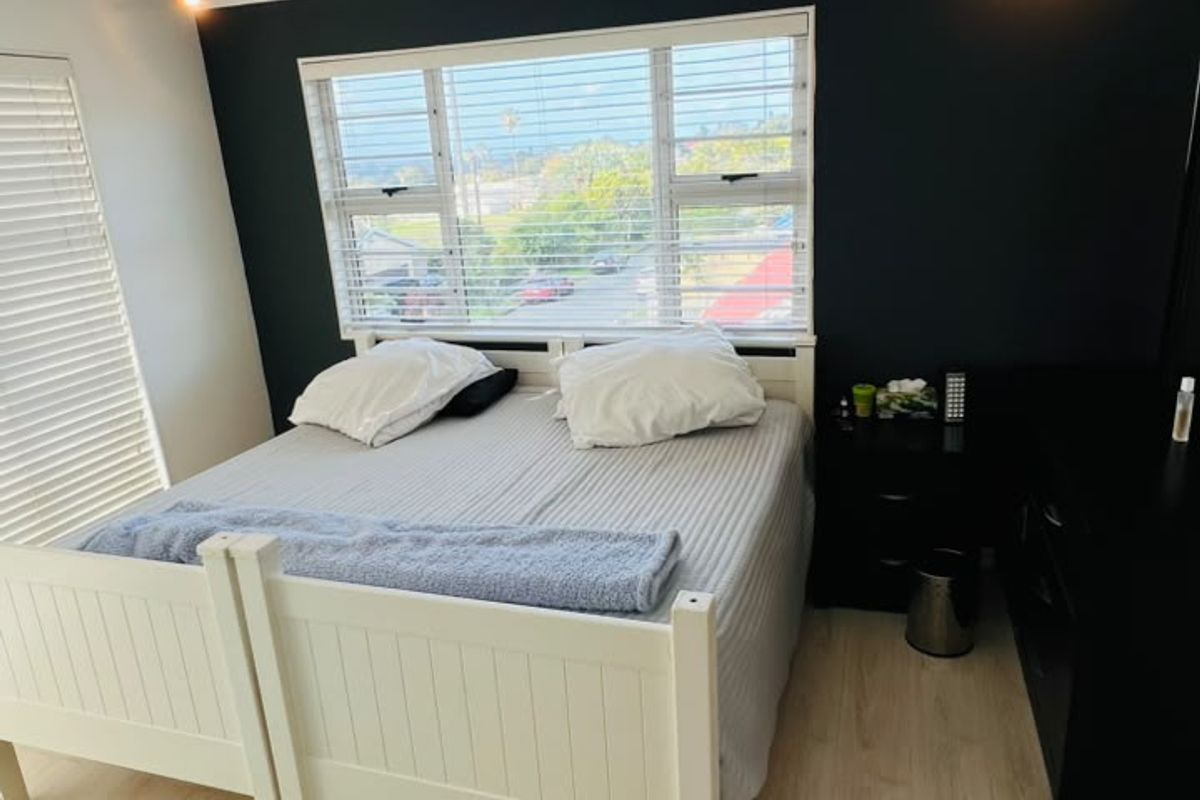 A well-lit bedroom featuring light laminate flooring, a dark accent wall, and large windows with venetian blinds offering an exterior view.