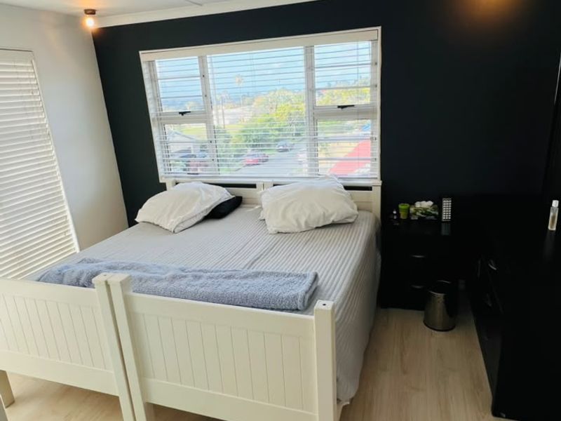 A well-lit bedroom featuring light laminate flooring, a dark accent wall, and large windows with venetian blinds offering an exterior view.