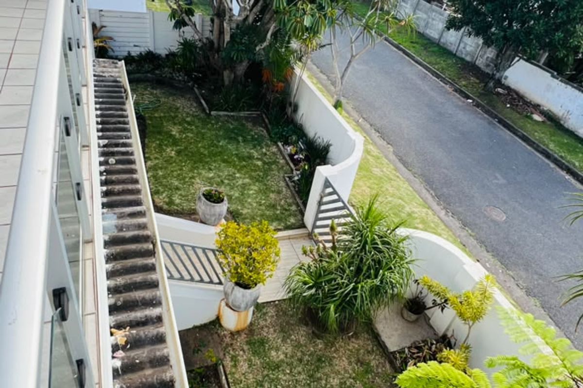 Elevated view from a balcony overlooking a landscaped garden, paved pathways, and retaining walls, with a view towards the adjacent street.