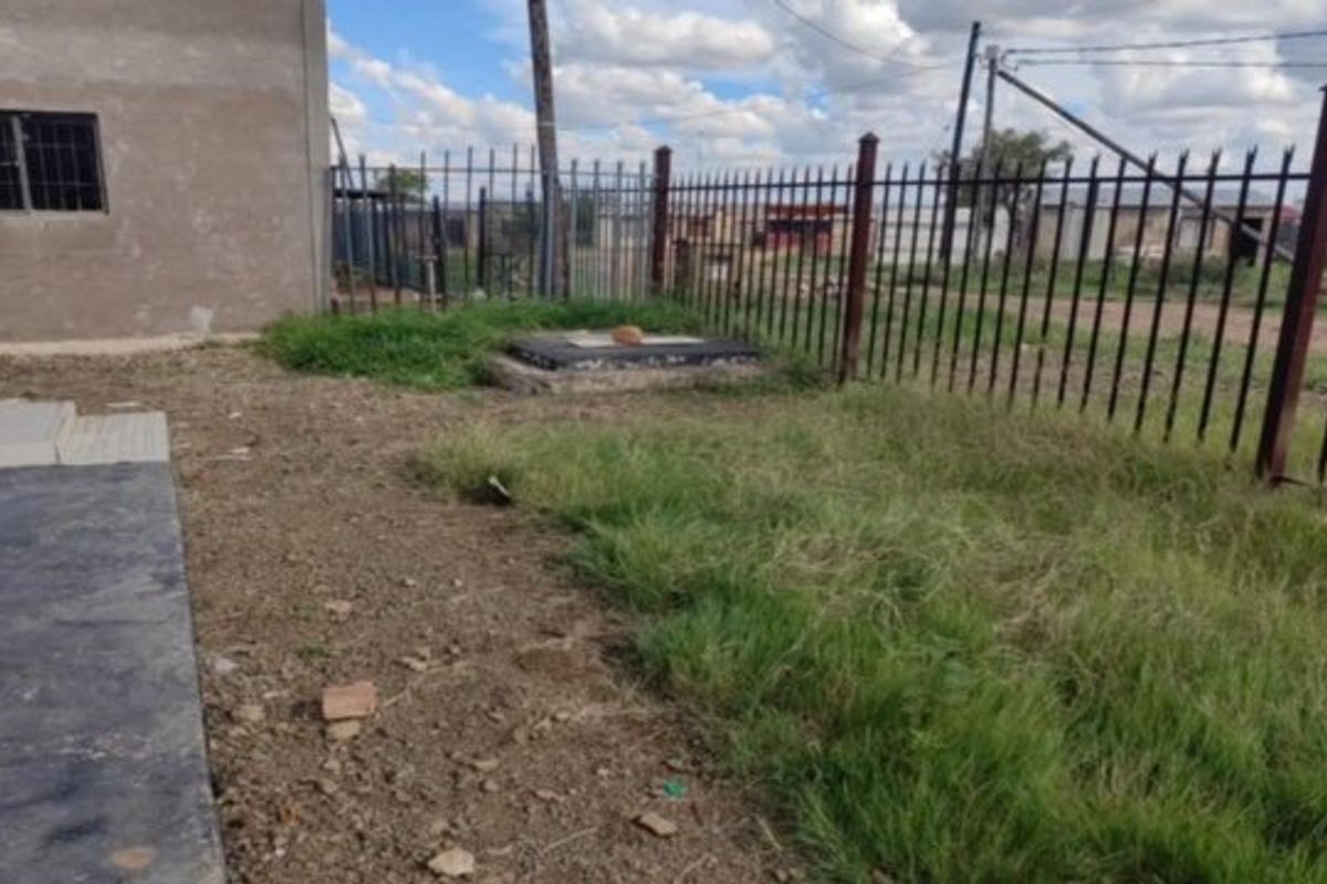 Rear exterior area featuring metal perimeter fencing, a concrete walkway, and an unpaved garden space with unfinished wall plaster.