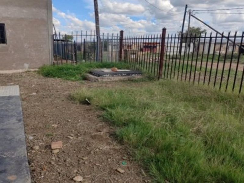 Rear exterior area featuring metal perimeter fencing, a concrete walkway, and an unpaved garden space with unfinished wall plaster.