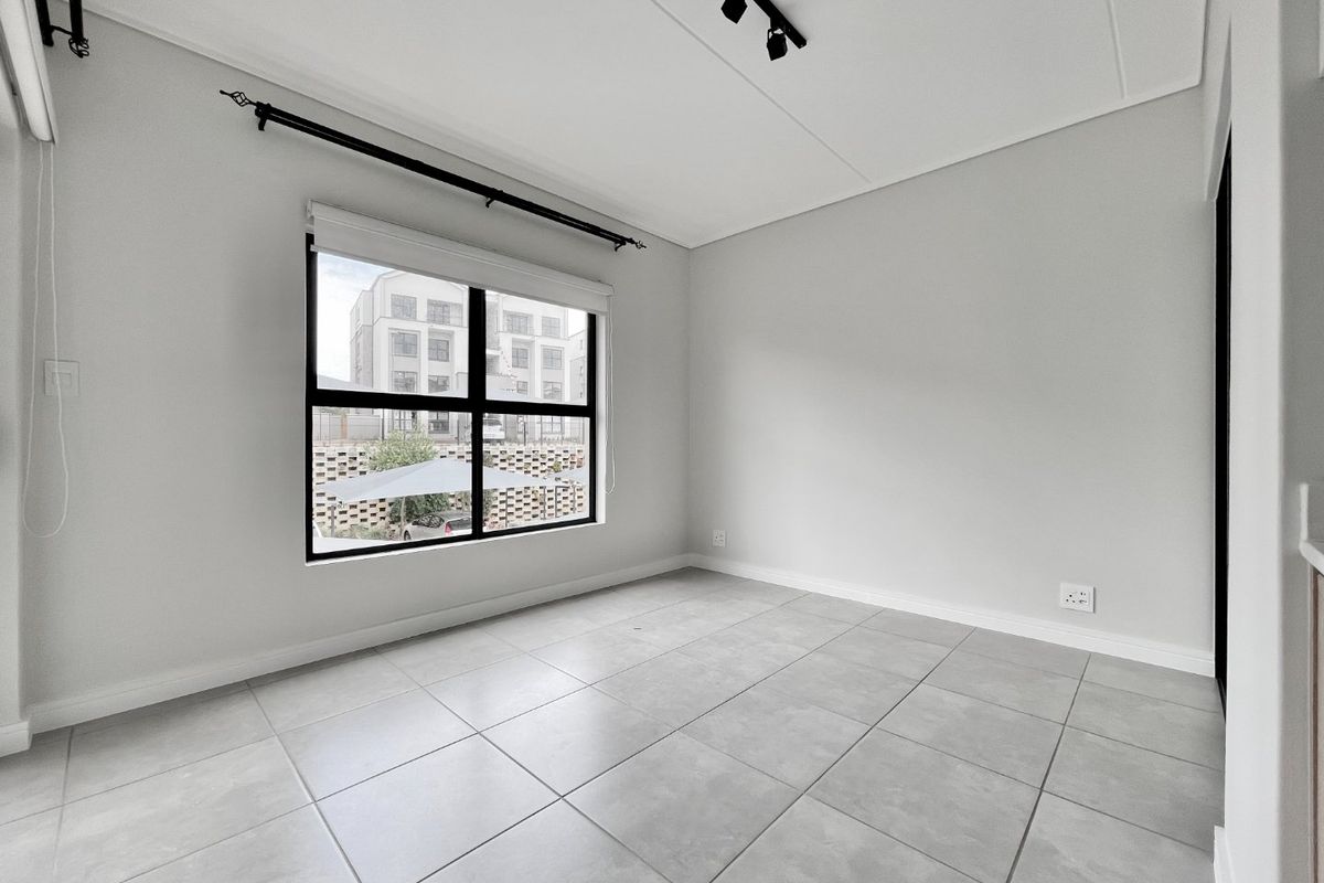A bright, empty bedroom featuring neutral tiled flooring, modern black-framed windows, and track lighting, providing a clean and versatile space ready for personalization.
