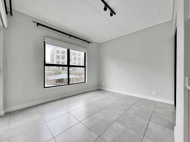 A bright, empty bedroom featuring neutral tiled flooring, modern black-framed windows, and track lighting, providing a clean and versatile space ready for personalization.