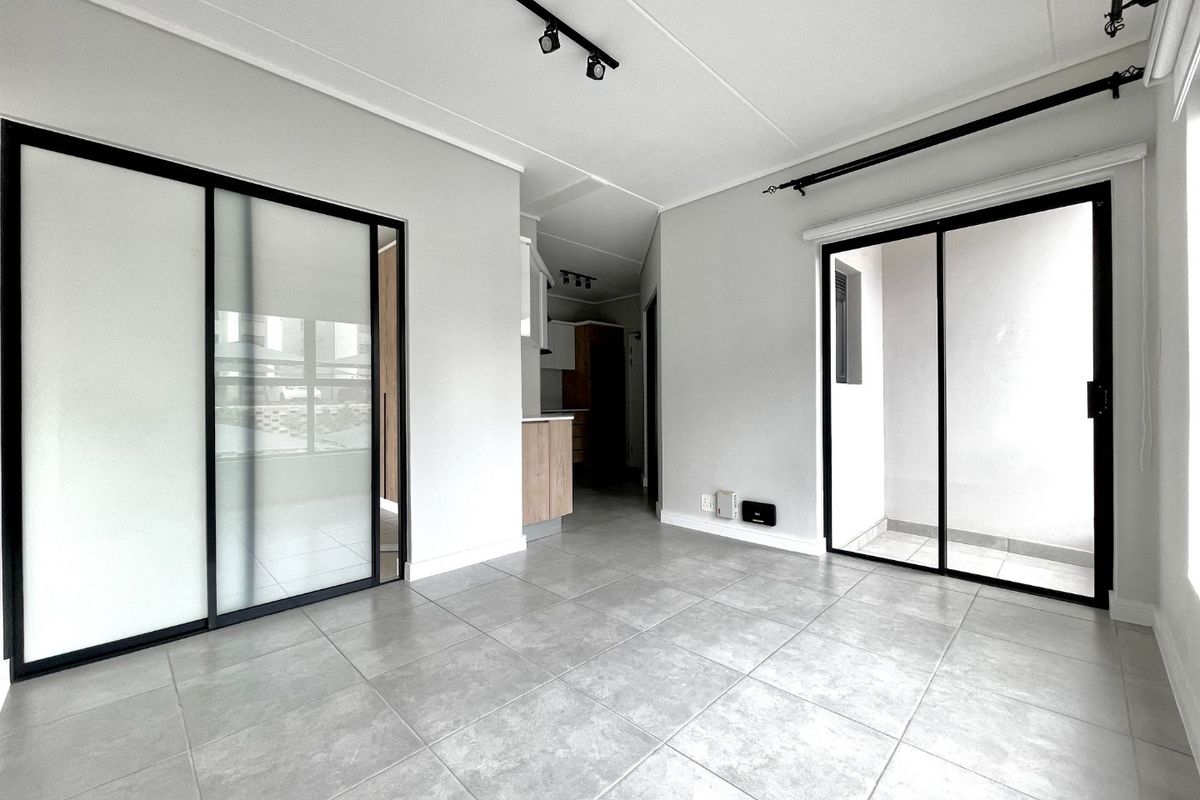 A bright, modern open-plan living area featuring neutral tiled flooring, sleek black-framed sliding doors, and contemporary track lighting.