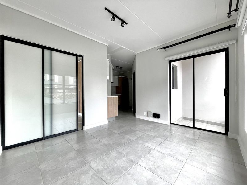 A bright, modern open-plan living area featuring neutral tiled flooring, sleek black-framed sliding doors, and contemporary track lighting.
