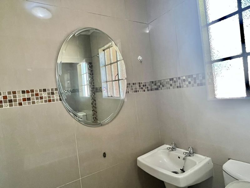 A functional bathroom featuring a pedestal basin, dual-handle mixer, oval mirror, and neutral wall tiling with a decorative mosaic border.