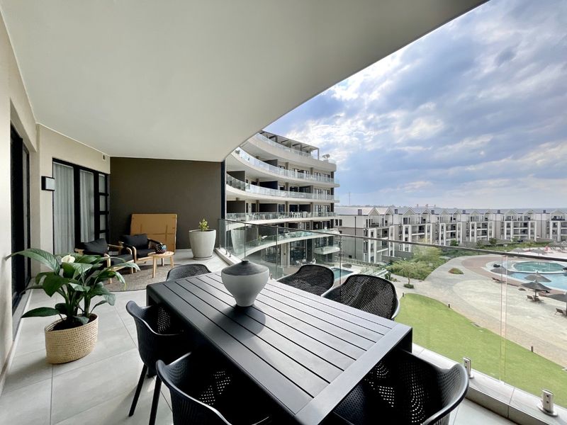 Spacious covered balcony featuring modern glass balustrades and tiled flooring, offering expansive views over the complex grounds and swimming pool area.