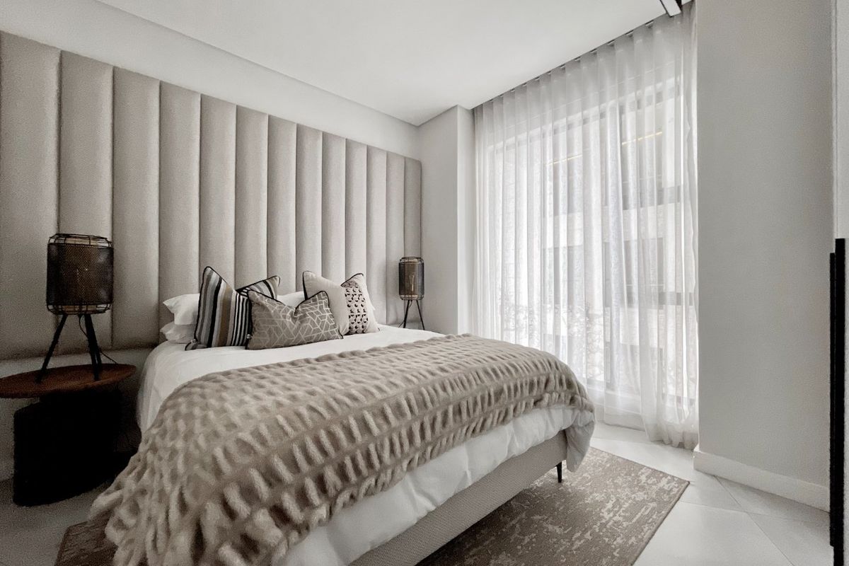 A modern, well-maintained bedroom featuring floor-to-ceiling upholstered headboard paneling, neutral tones, and large windows with elegant sheer curtains.