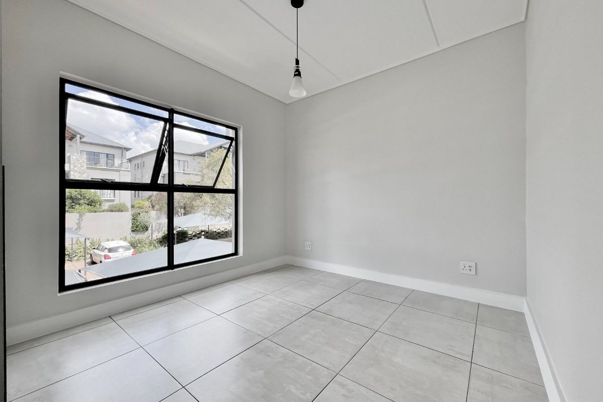 A bright, empty bedroom featuring modern grey floor tiles, neutral walls, and a large black-framed window providing natural light.