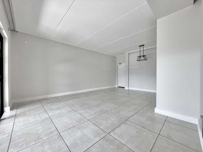 A bright, open-plan living area featuring neutral grey floor tiles, clean white walls, and a modern pendant light fixture.