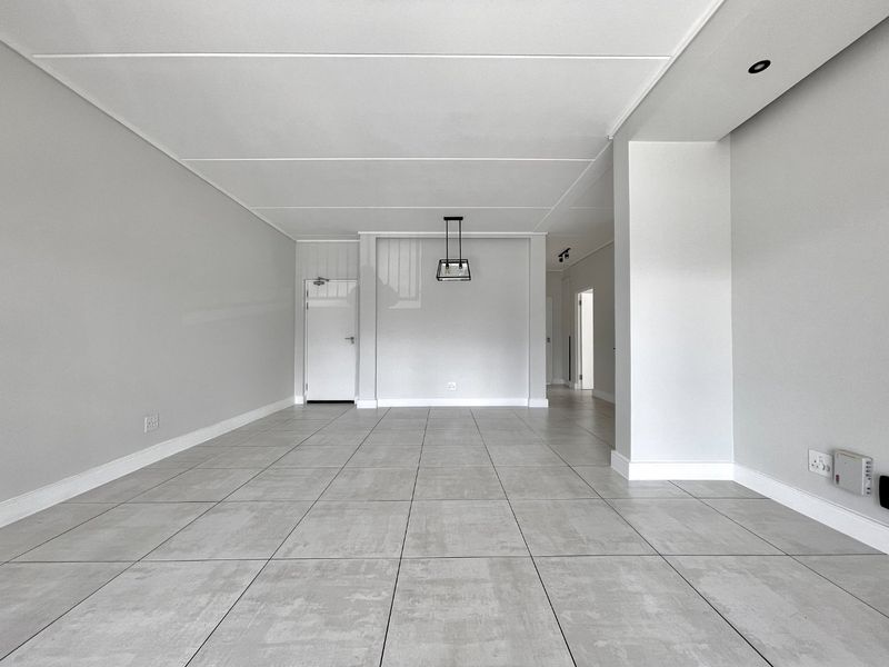 A spacious, empty living area featuring neutral grey floor tiles, clean white walls, and modern recessed and pendant lighting.