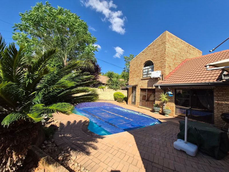 A well-maintained outdoor pool area featuring a swimming pool with a cover, surrounded by brick paving and a face brick house exterior.