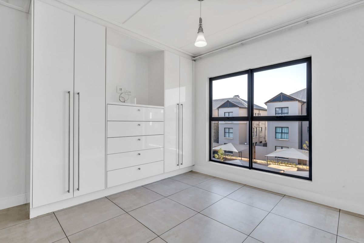 A bright, empty bedroom featuring modern white built-in cupboards, large windows with neighbourhood views, and neutral tiled flooring.