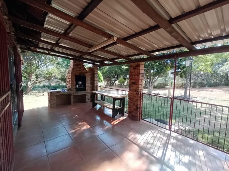 Covered outdoor patio featuring a built-in brick braai, tiled flooring, and exposed timber beams, overlooking the garden area.