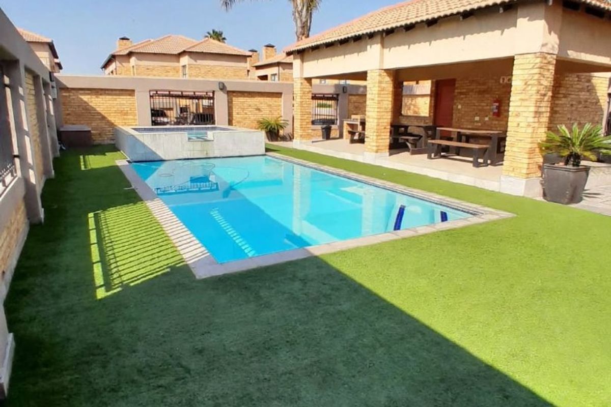 A well-maintained outdoor pool area featuring a swimming pool, artificial lawn, and a covered patio structure with brick detailing.