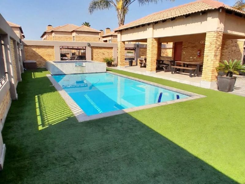 A well-maintained outdoor pool area featuring a swimming pool, artificial lawn, and a covered patio structure with brick detailing.