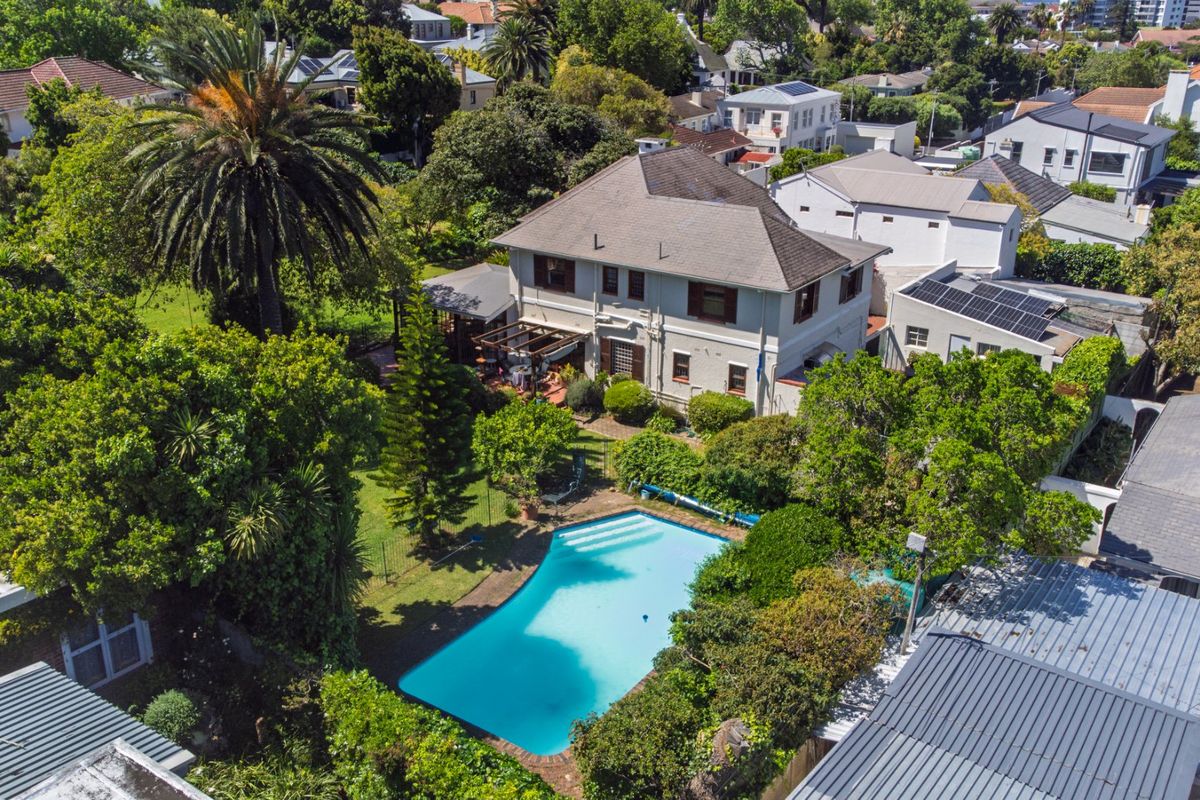 Aerial view of a residential property featuring a large swimming pool, established garden, covered patio area, and a multi-story house with a tiled roof.