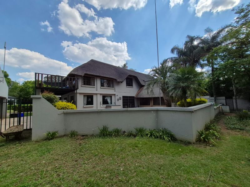 Rear view of a two-story house featuring a traditional thatch roof, a raised wooden deck, and a surrounding perimeter wall with a landscaped garden.