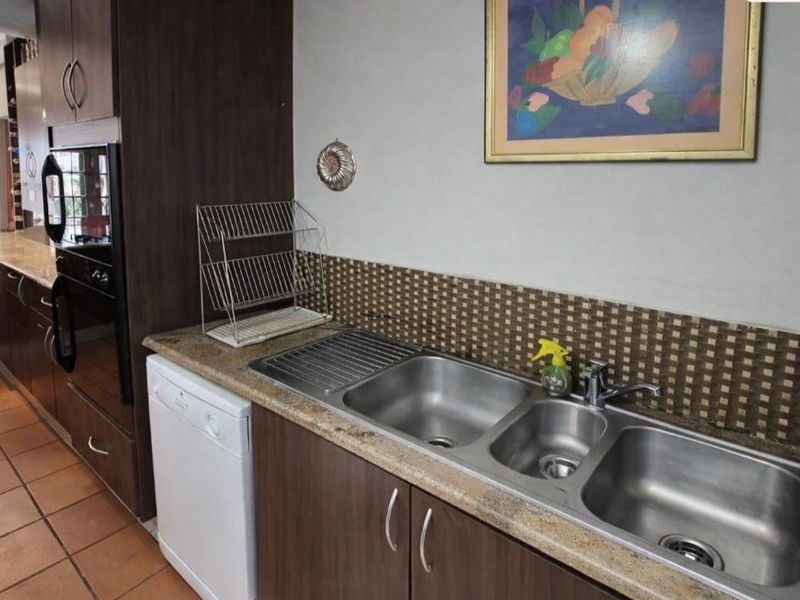 A functional kitchen featuring dark wood cabinetry, stone countertops, a triple stainless steel sink, and a mosaic tile backsplash with ceramic floor tiles.